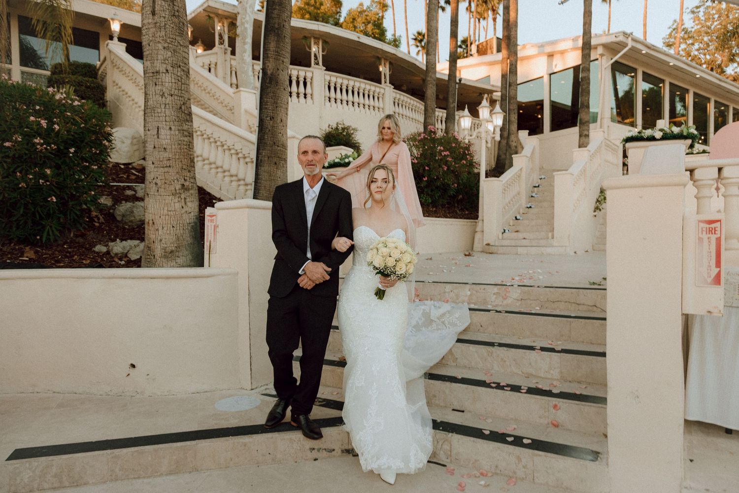 A wedding couple posing on white concrete steps with tropical palm trees and string lights in the background.