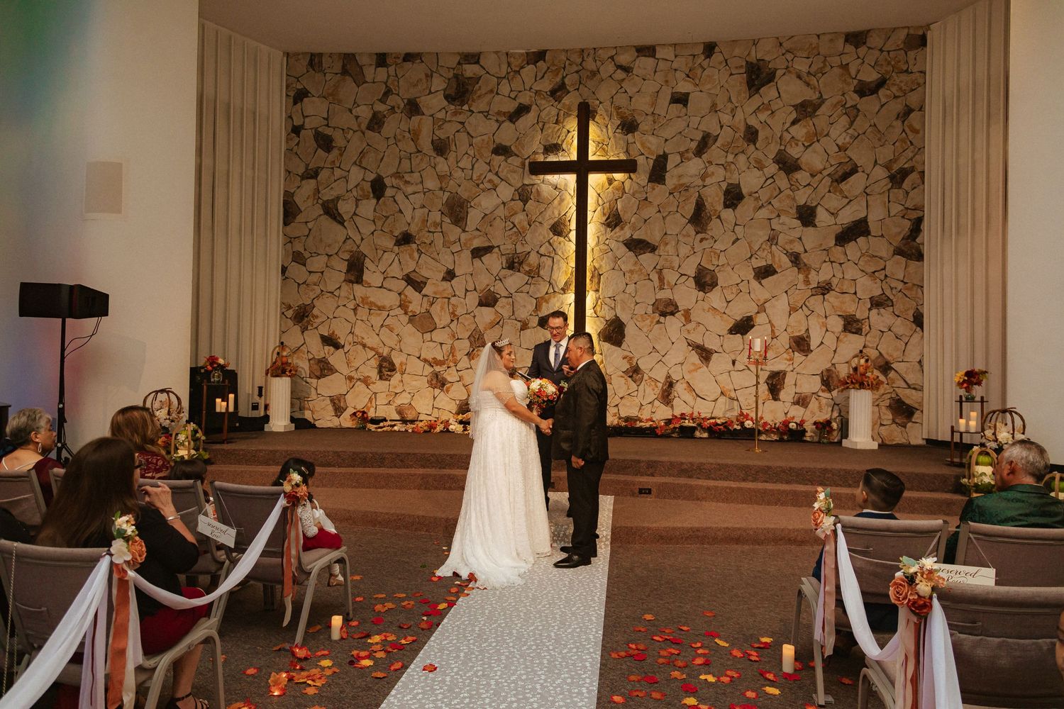 Wedding ceremony at church altar with stone wall backdrop and red rose petals lining white aisle runner.