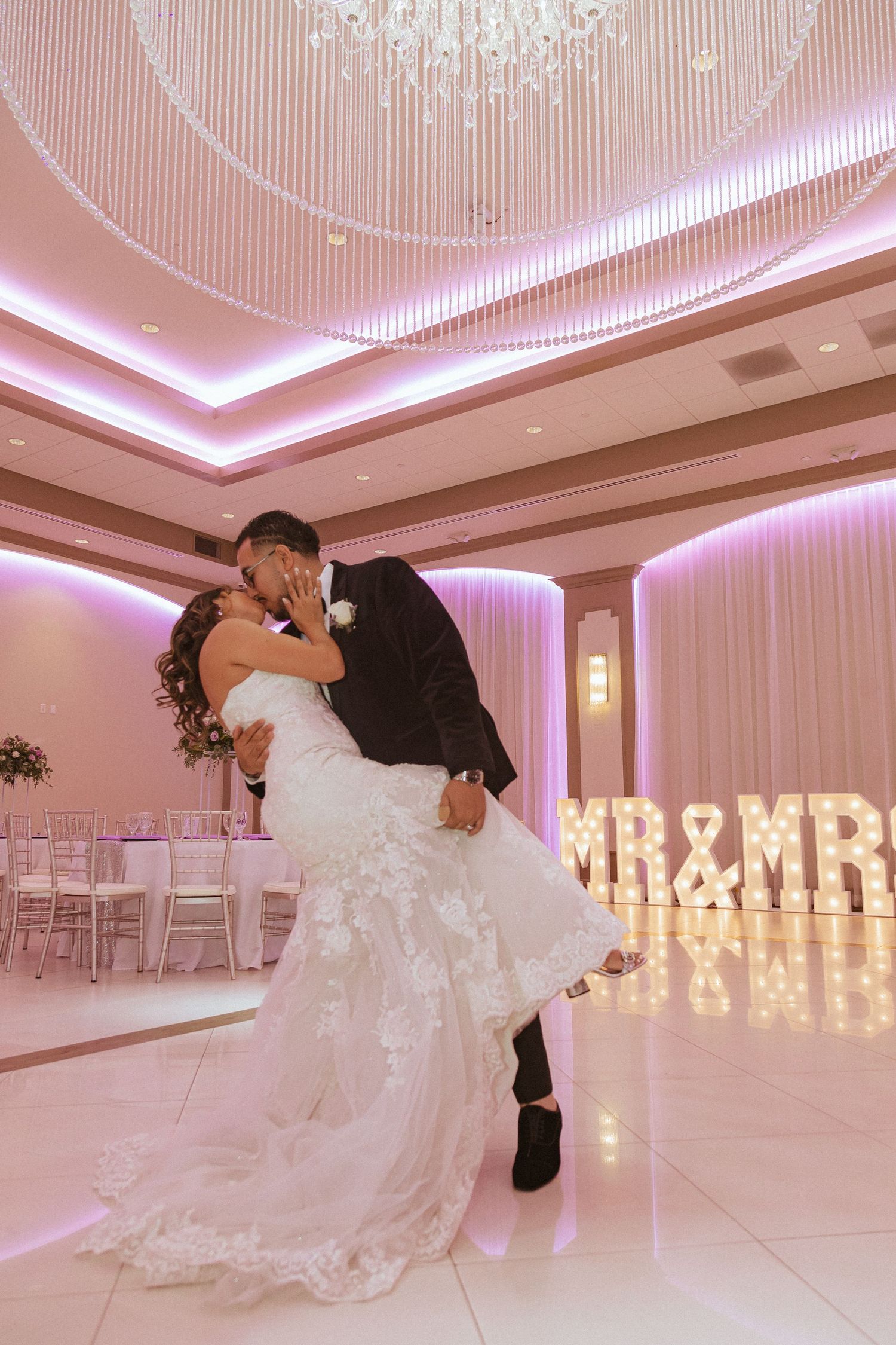 Couple performs romantic dip pose in front of glowing MR & MRS marquee letters at their wedding reception.