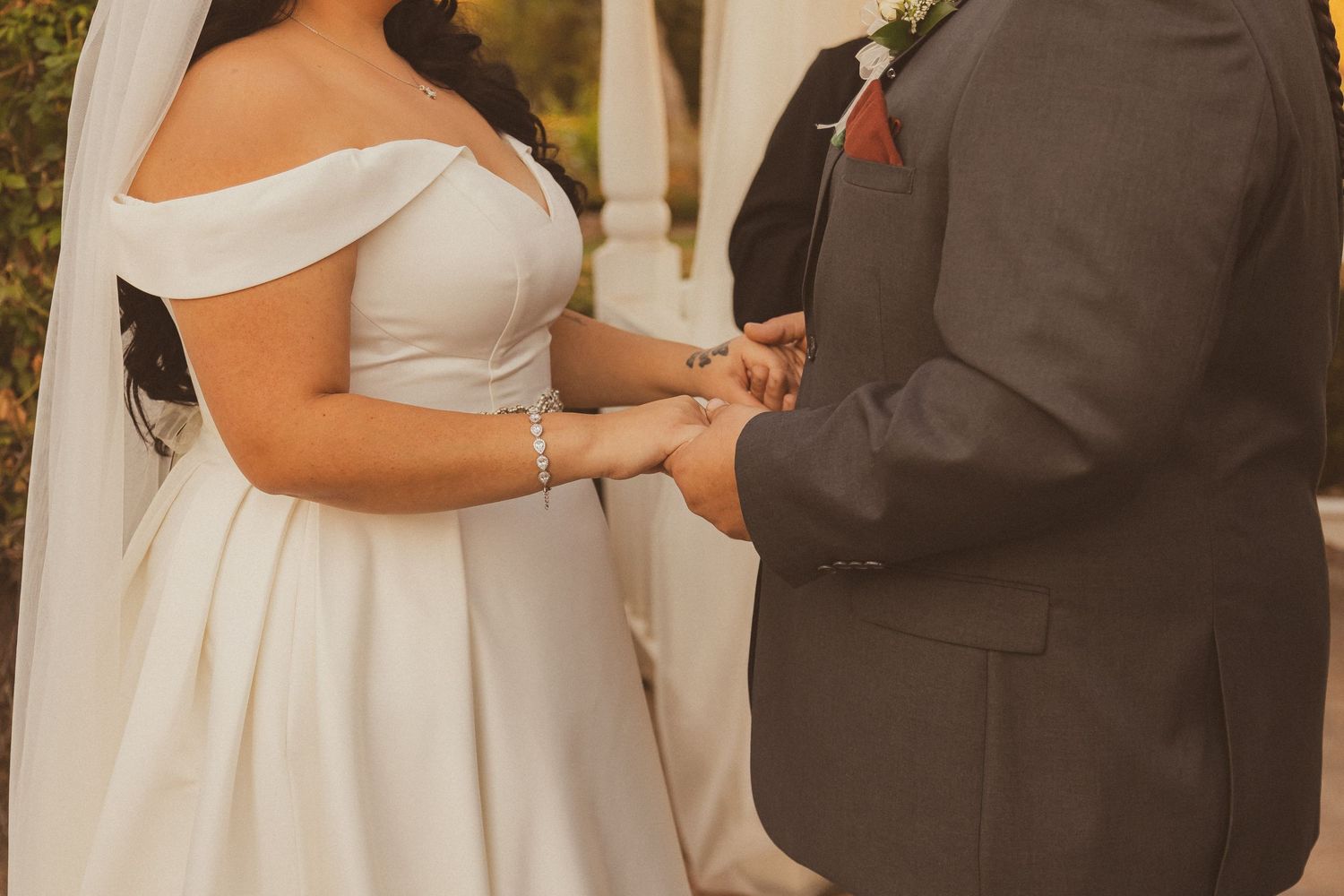Close-up of bride in off-shoulder gown exchanging rings with groom during outdoor ceremony.