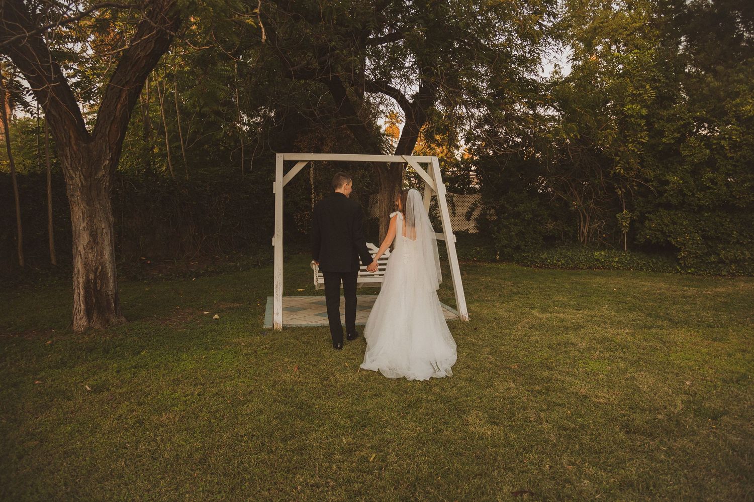 Newlyweds stand together under wooden arch in outdoor garden setting at sunset.