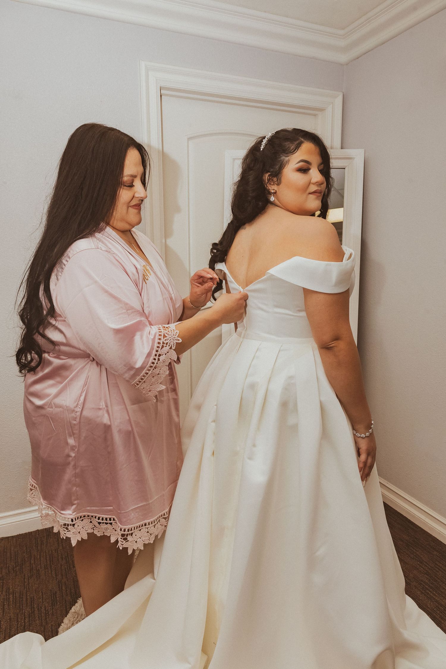 Final adjustments being made to an elegant white off-shoulder wedding gown with pleated skirt.