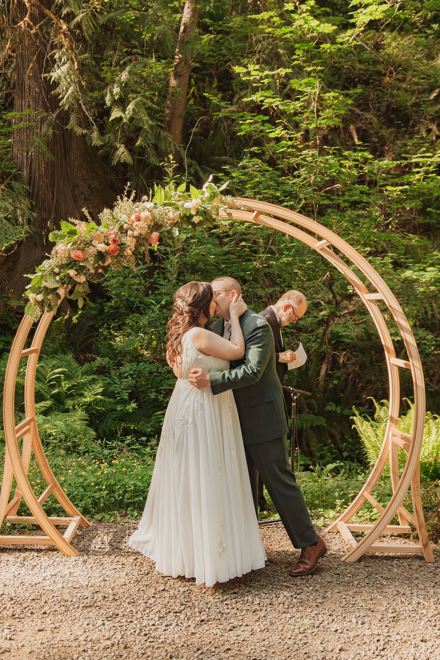 A romantic wedding moment under a floral arch in a forest setting.