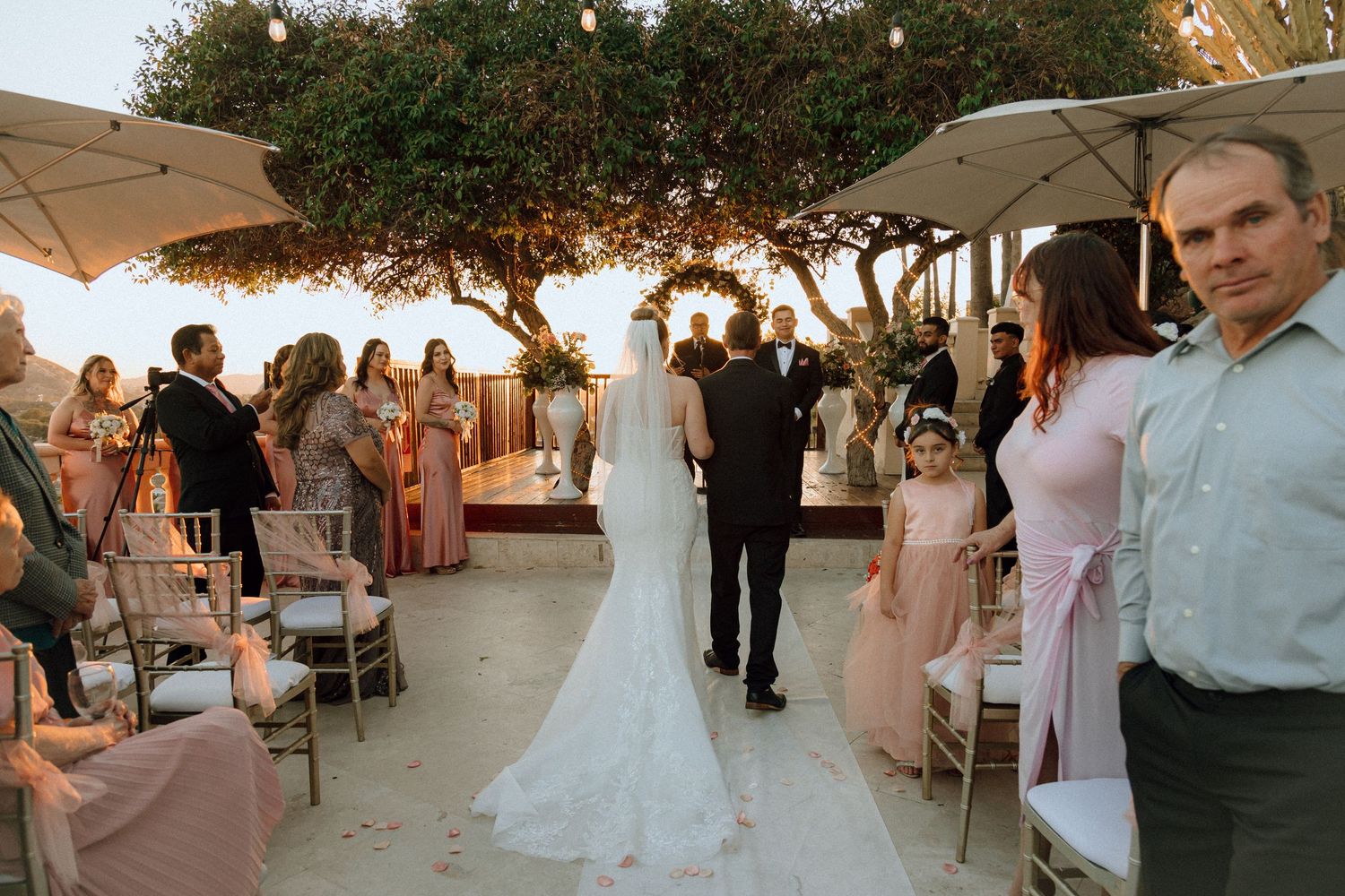 Guests watch a wedding ceremony procession under string lights and umbrellas at dusk.