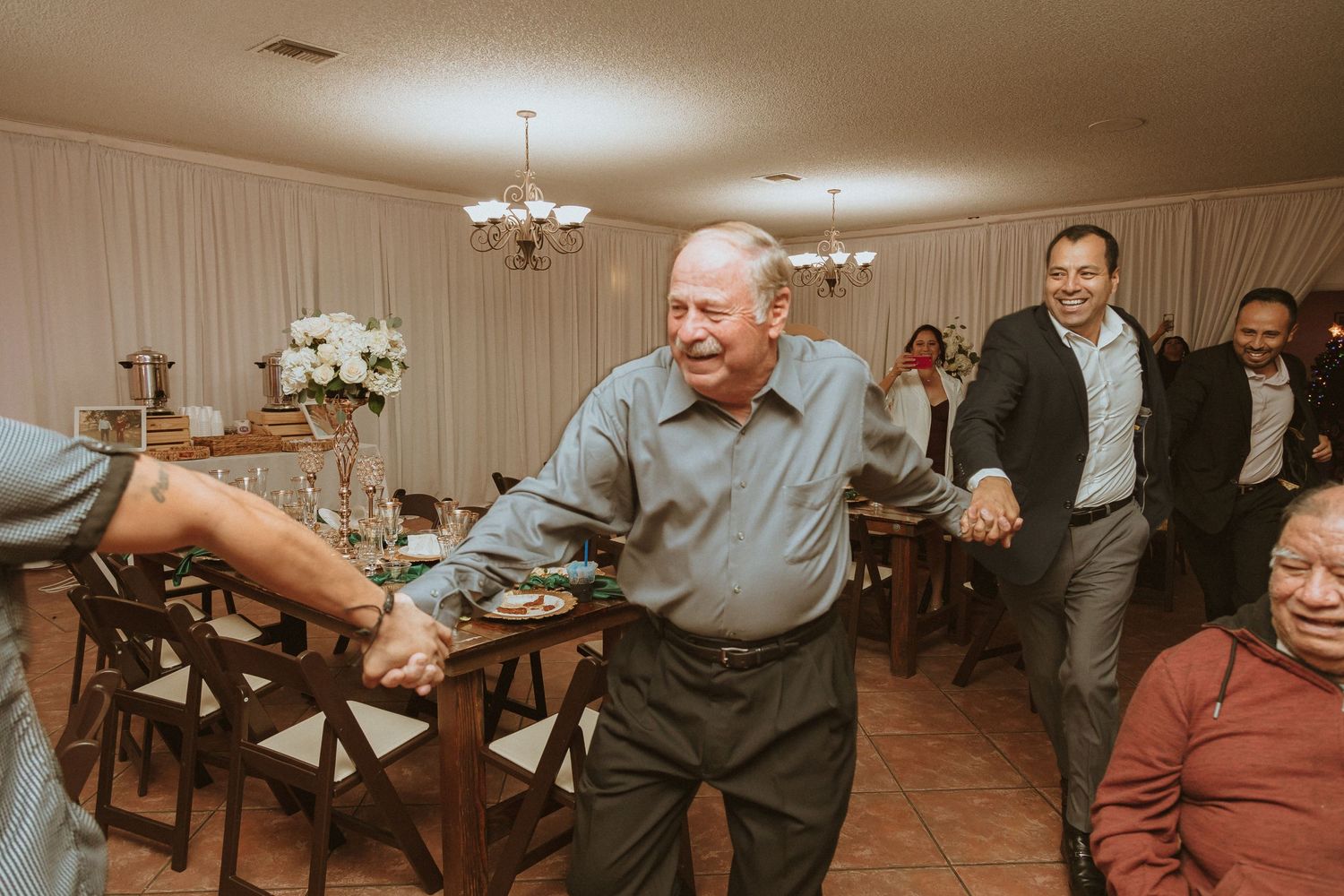 Wedding guests dance in formation on a tiled floor during an indoor reception.