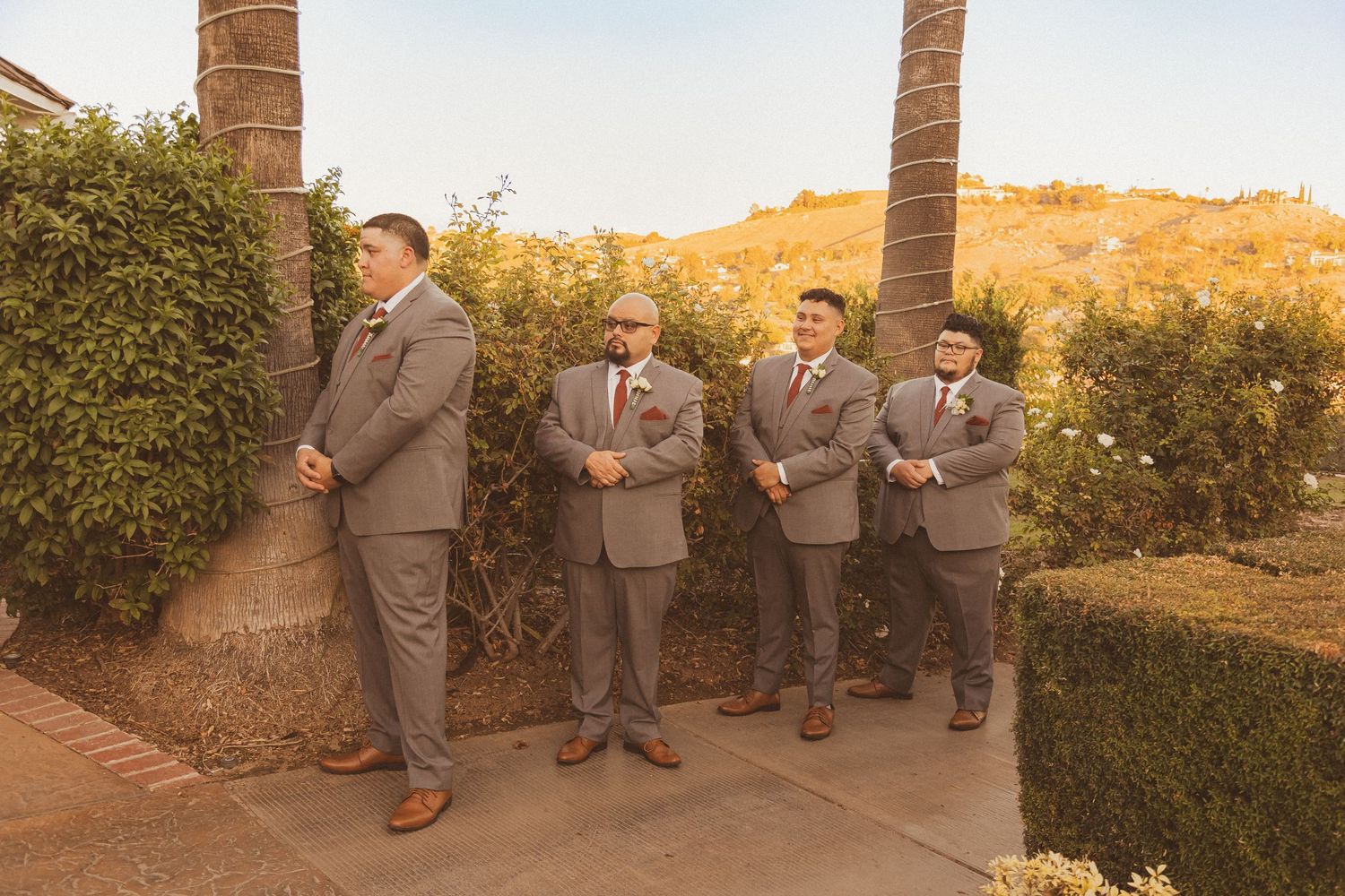 Four groomsmen in grey suits pose beside palm trees during sunset.