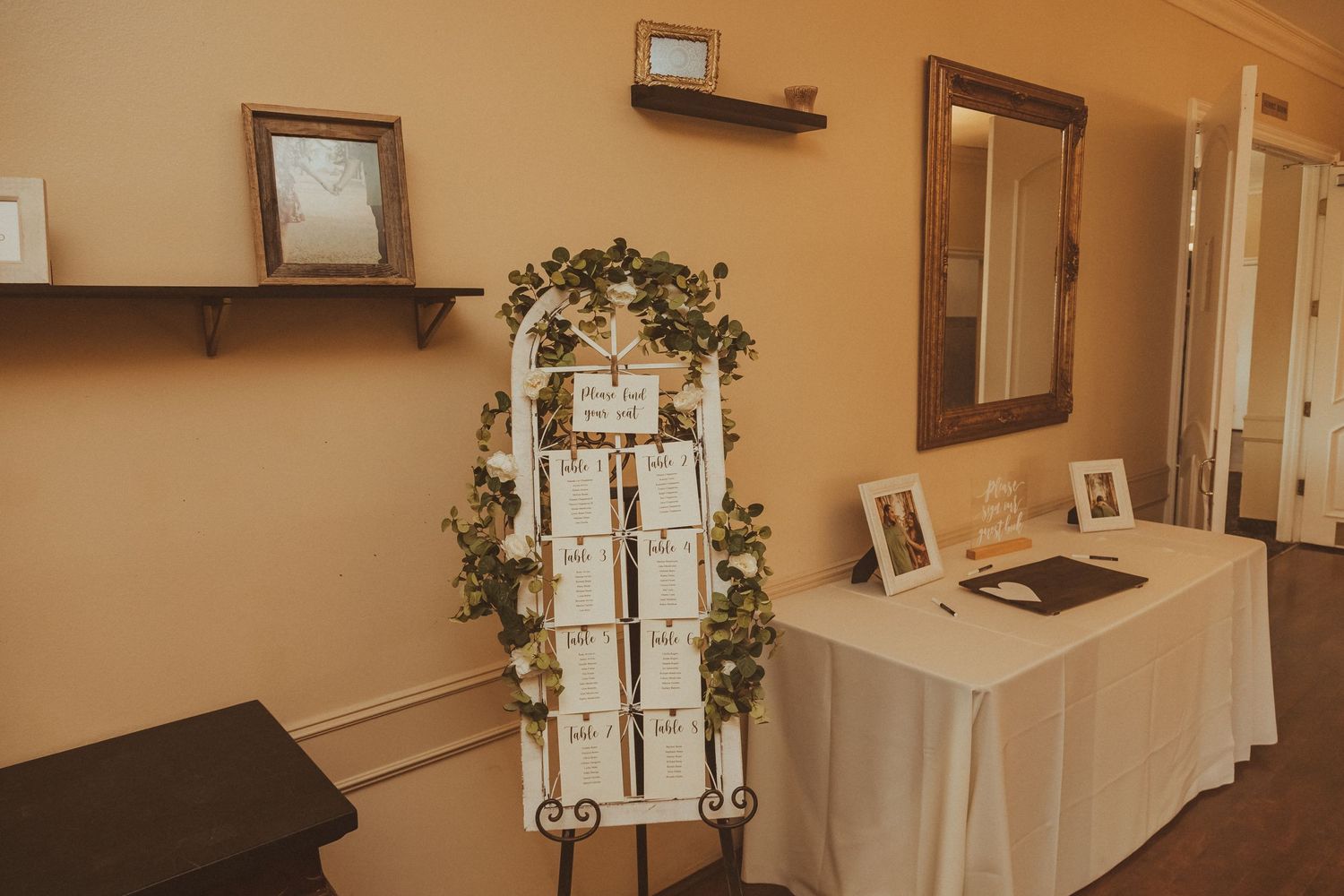 A wedding seating chart displayed on an easel decorated with greenery and framed by photo displays.