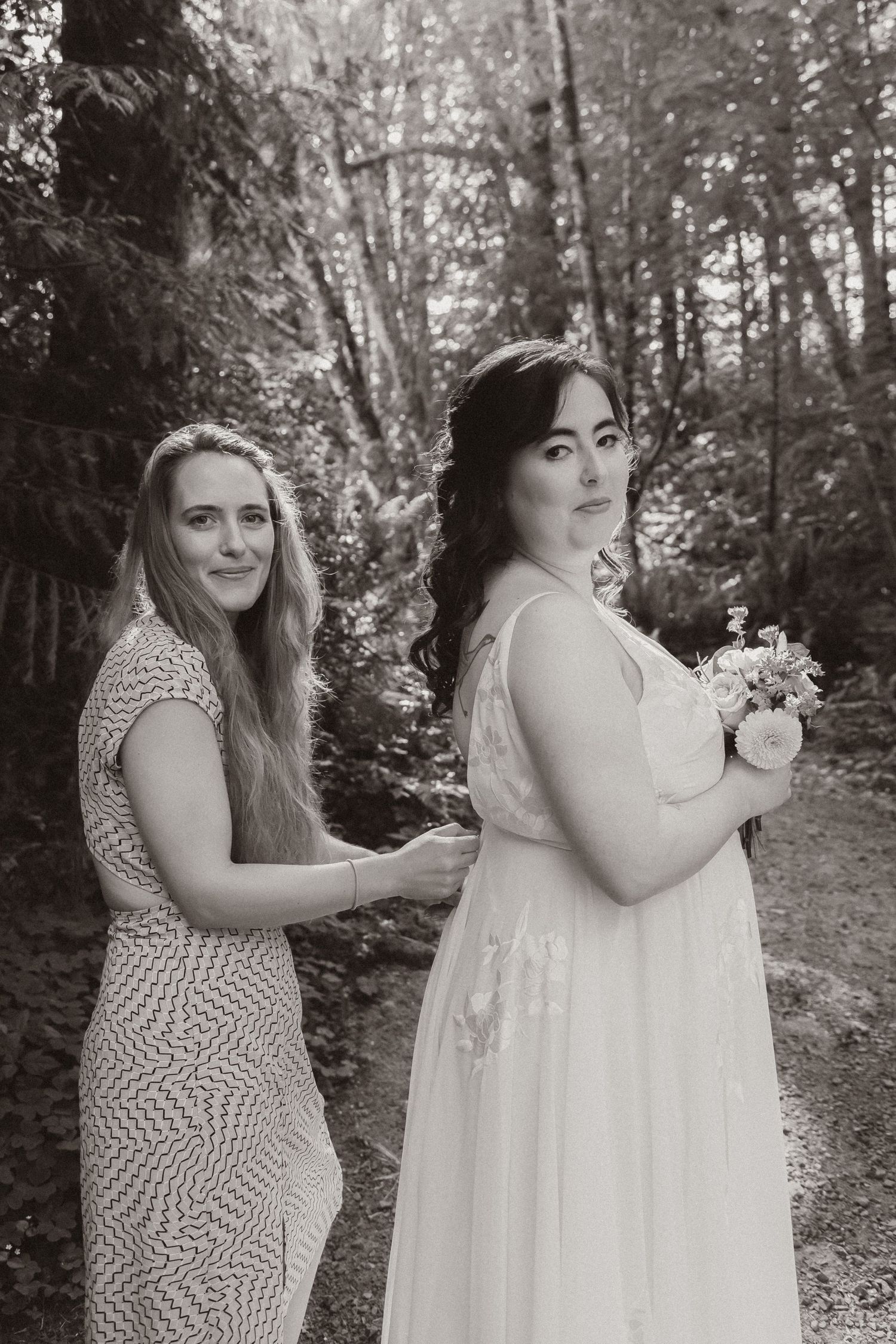 Two women pose in formal dresses in a forest setting, one wearing a patterned gown and one in white.