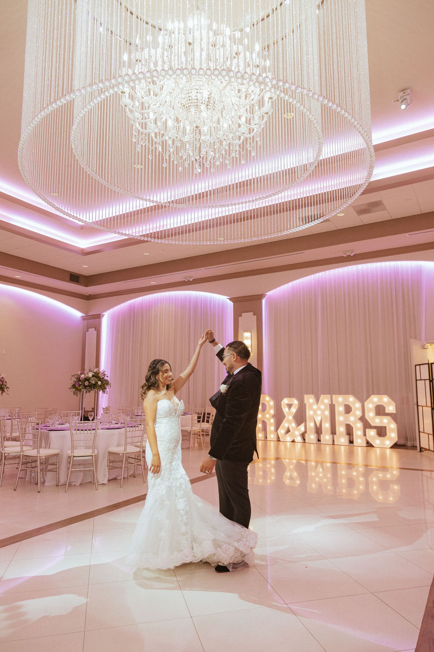 Wedding couple dancing in a pink-lit ballroom beneath a grand crystal chandelier.