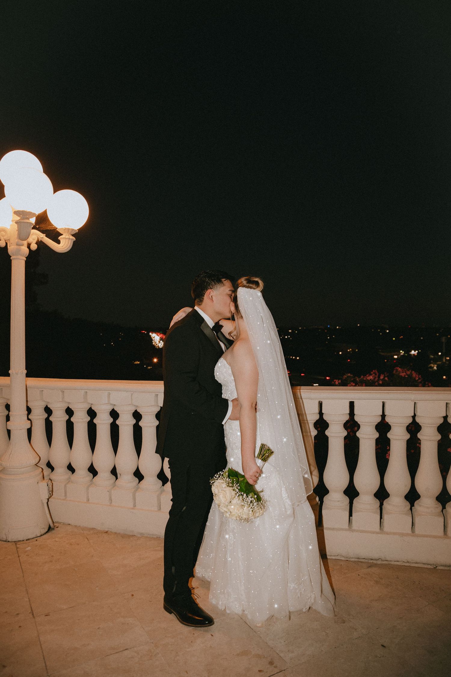 Newlyweds share a romantic moment on a lit balcony overlooking city lights.