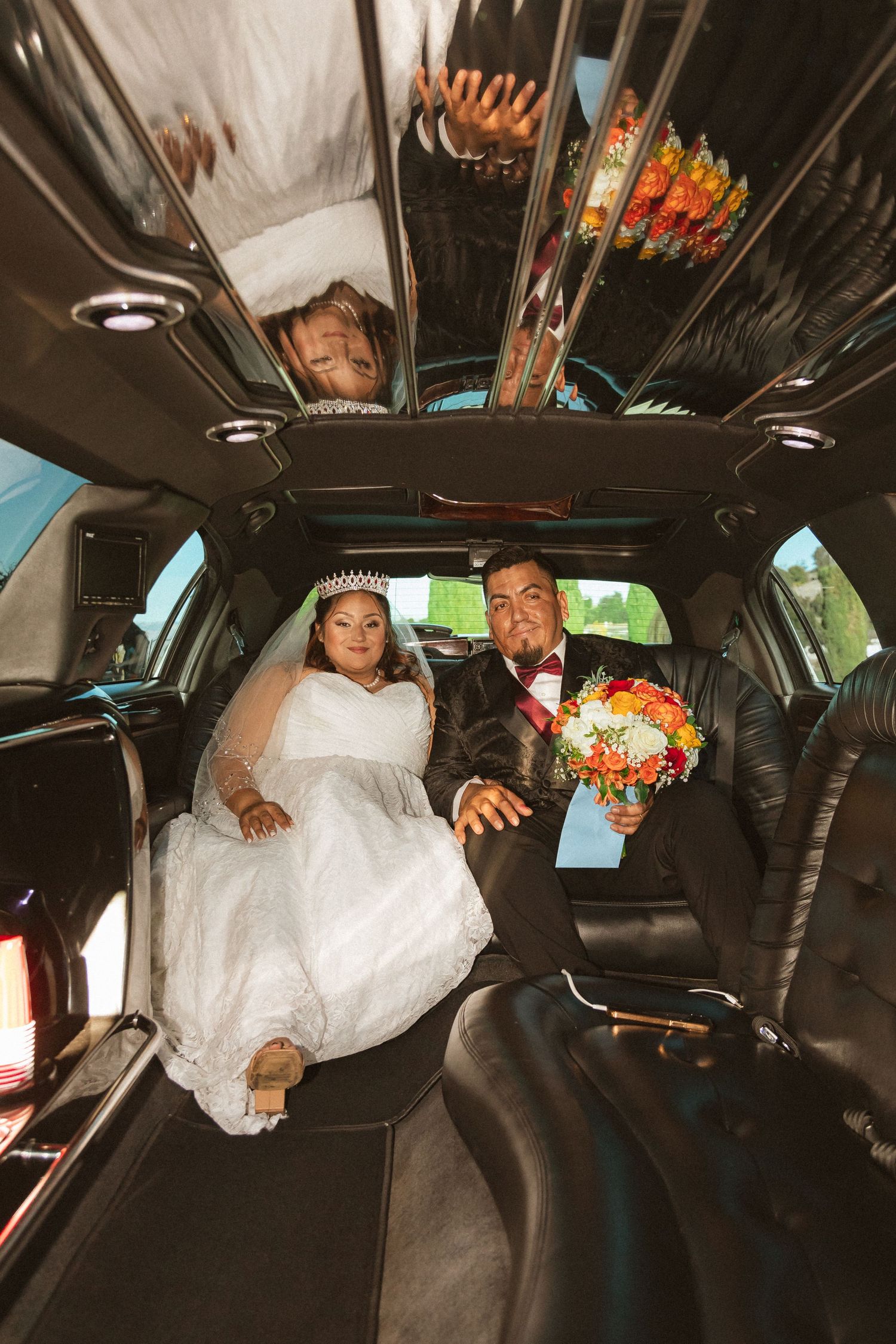 Wedding couple sitting in the luxurious black leather interior of a limousine with orange floral bouquet.