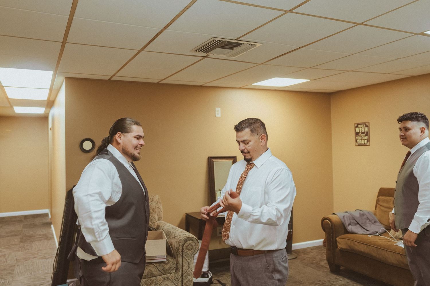 Three formally dressed people interact in a room with beige walls and fluorescent lighting.