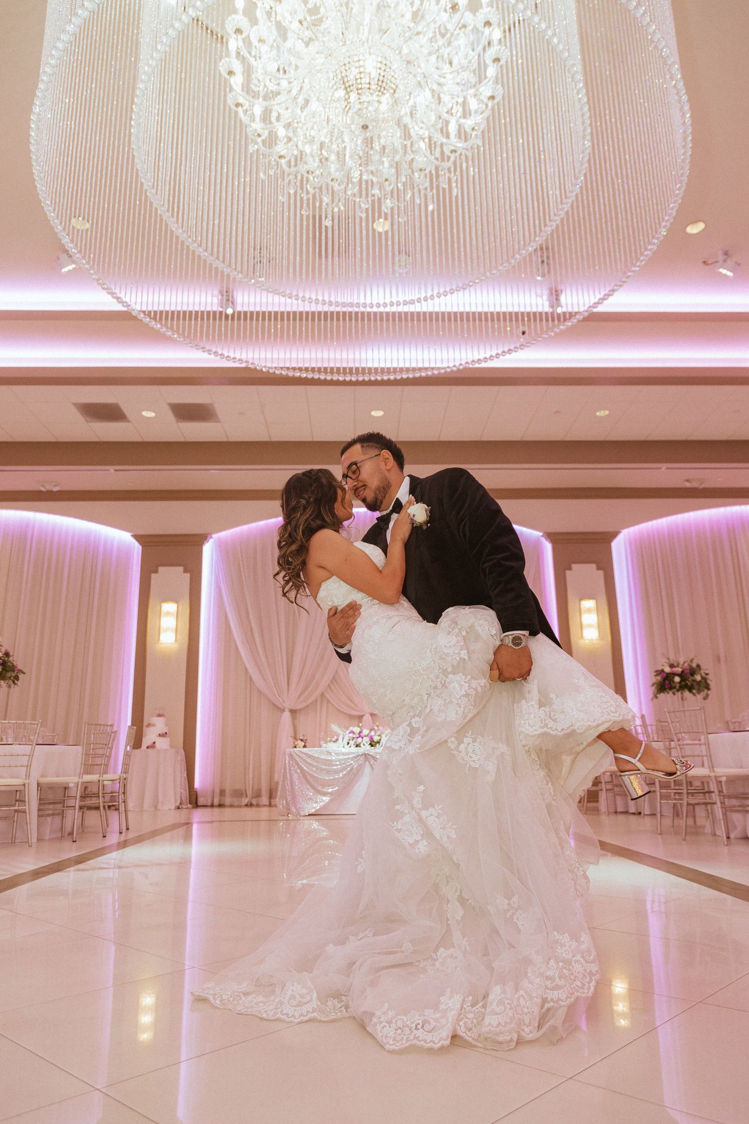 Dramatic ballroom dance dip under ornate chandelier with pink ambient lighting and white decor.