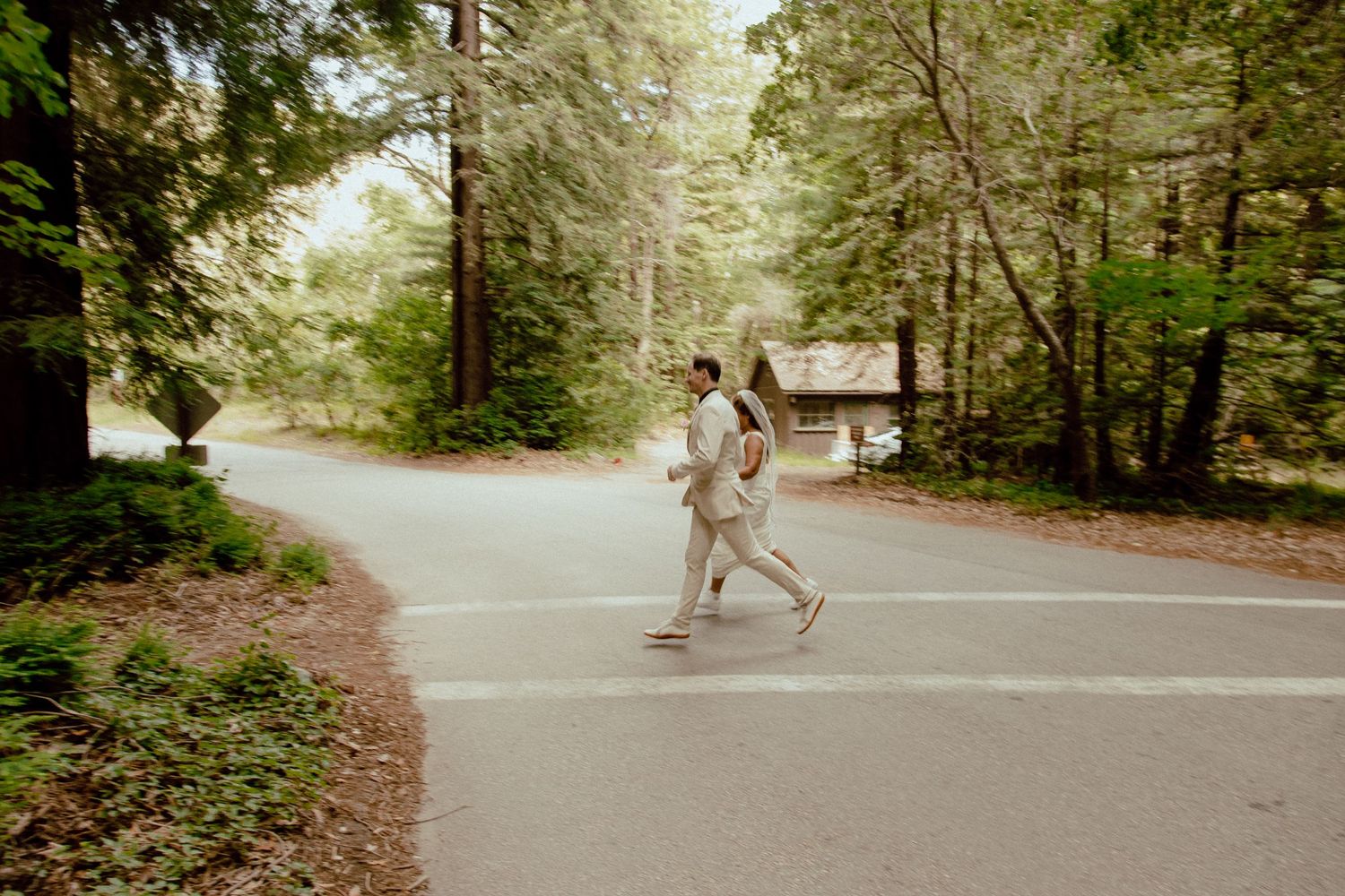 Two figures in light colored clothing run playfully down a forest road.
