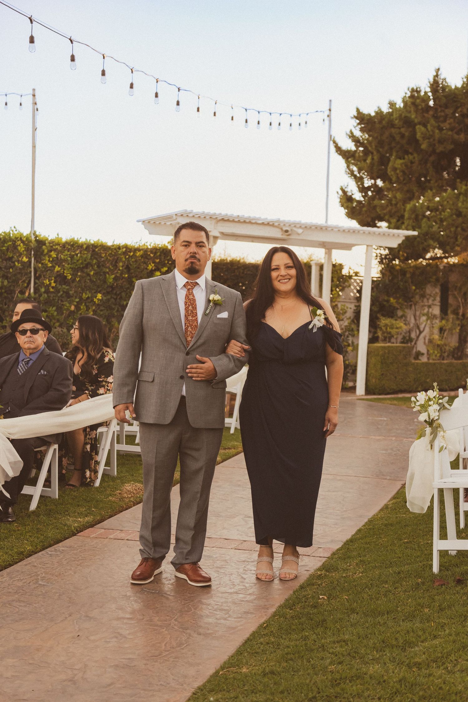 Wedding party members walk down an outdoor aisle with string lights overhead.