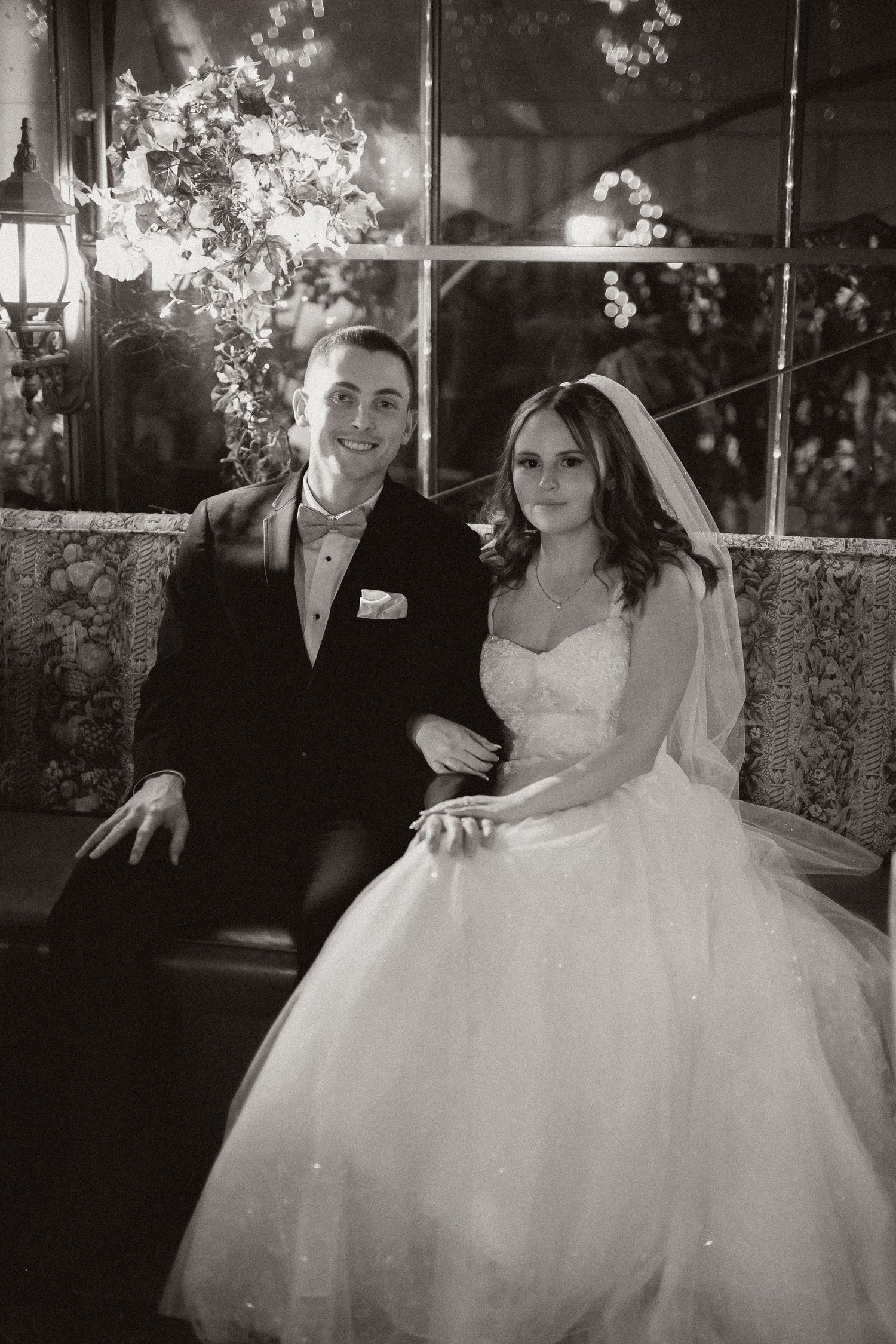Black and white wedding portrait of couple seated beneath elegant floral arrangements.