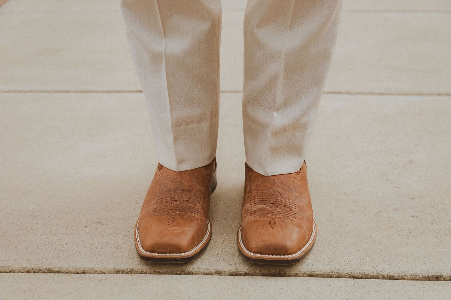 Brown leather dress shoes paired with white suit pants on a concrete floor.