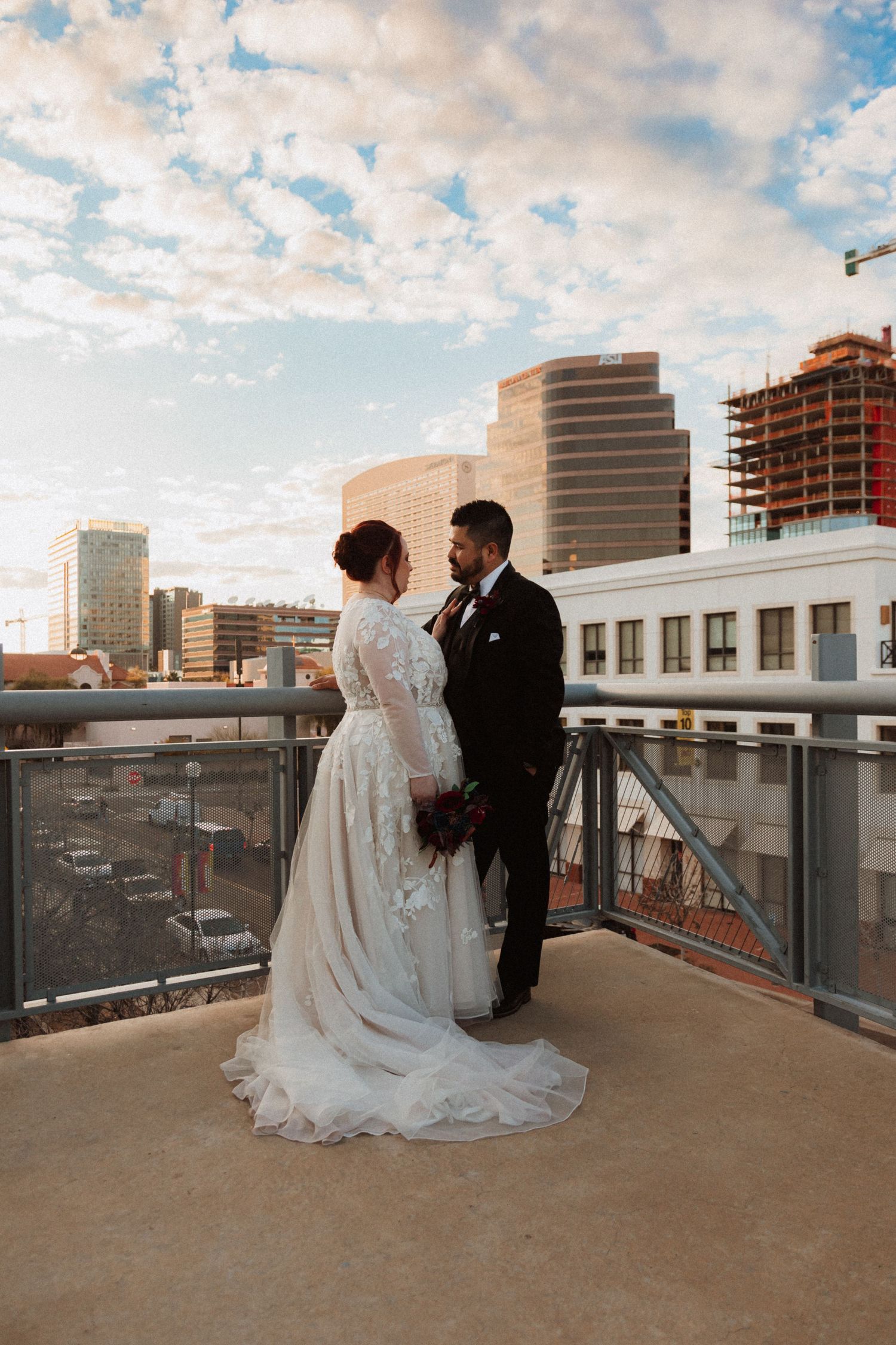 Couple in wedding attire pose on urban rooftop with downtown buildings and sunset sky.