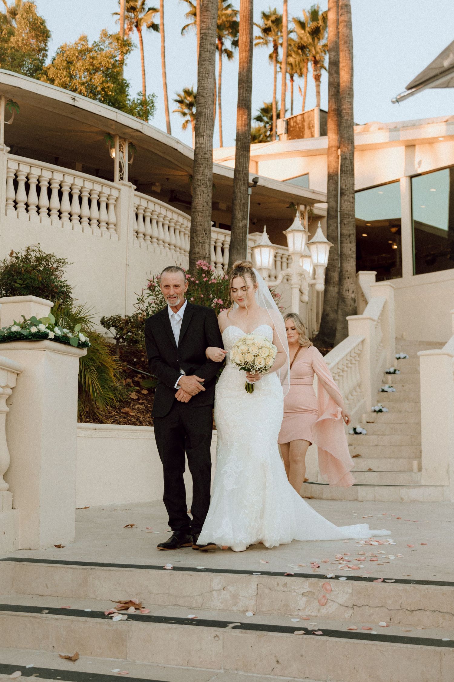 Wedding party poses on ornate white staircase with tall palm trees in California setting.