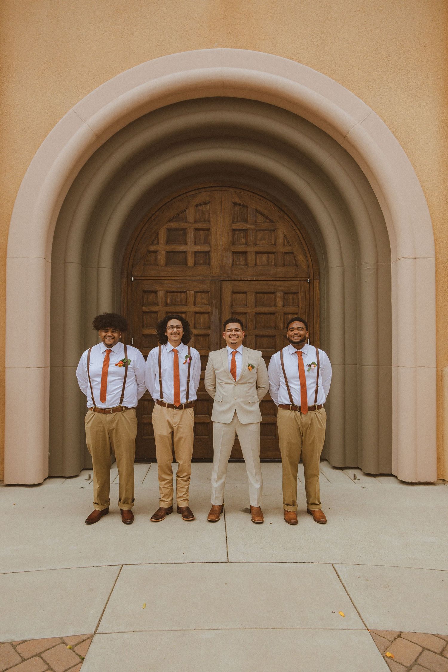 Groomsmen in khaki pants and orange suspenders pose for a wedding photo in front of an arched wooden door.