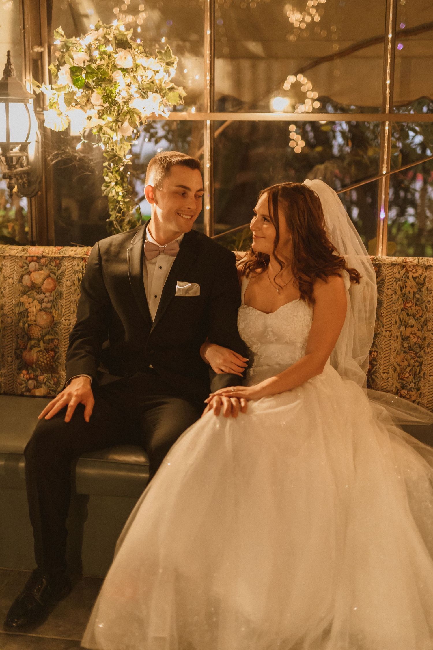 Romantic couple sits together in warm lighting with bride in white gown and groom in black tuxedo.