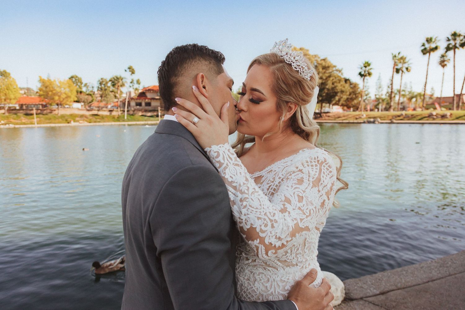 Newlyweds share an intimate kiss by a scenic lake with palm trees at sunset.