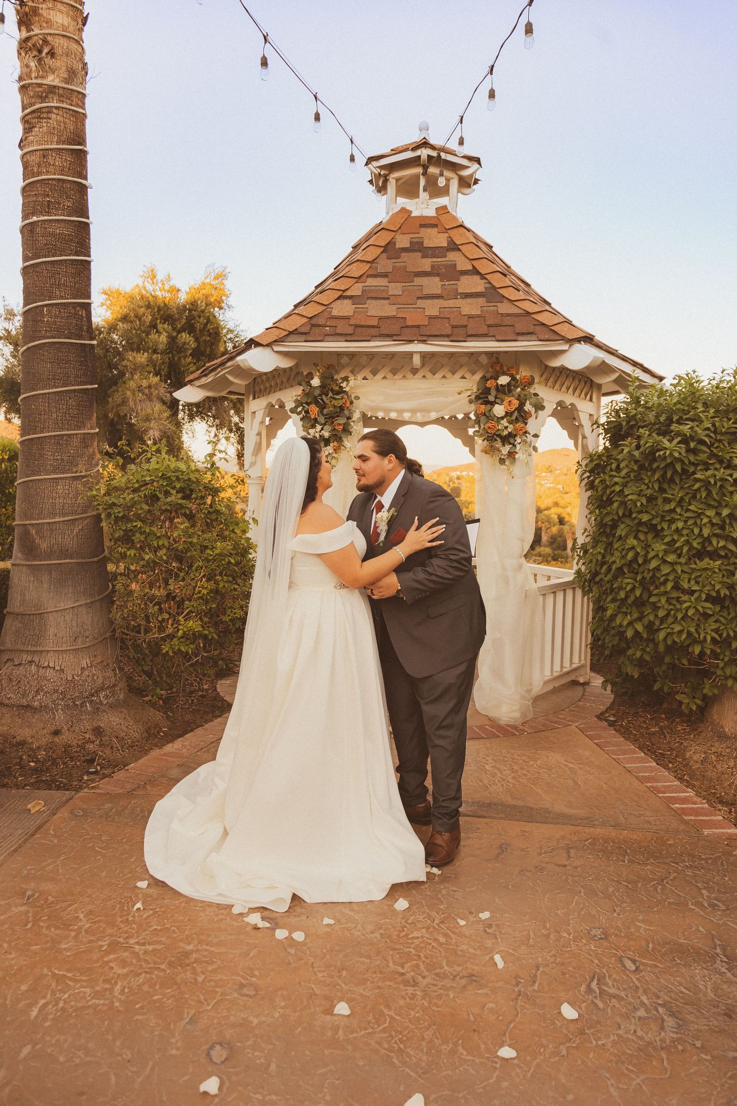 Newlyweds share an intimate moment under a wooden gazebo with string lights at sunset.