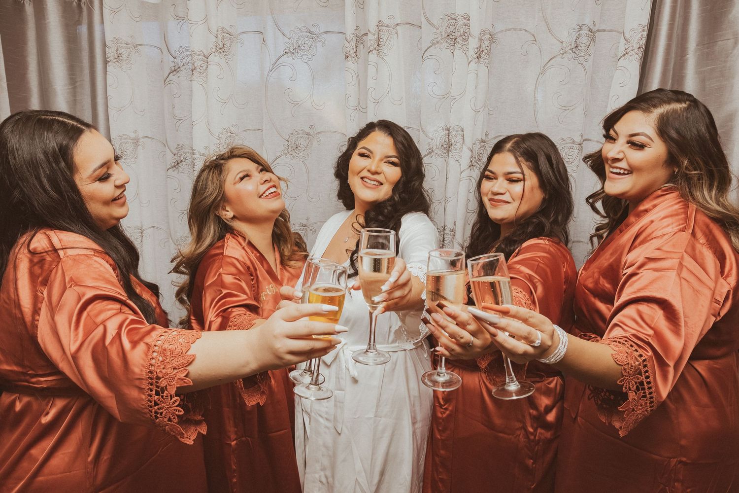 Bridal group in rust-colored robes celebrates with champagne against neutral backdrop.