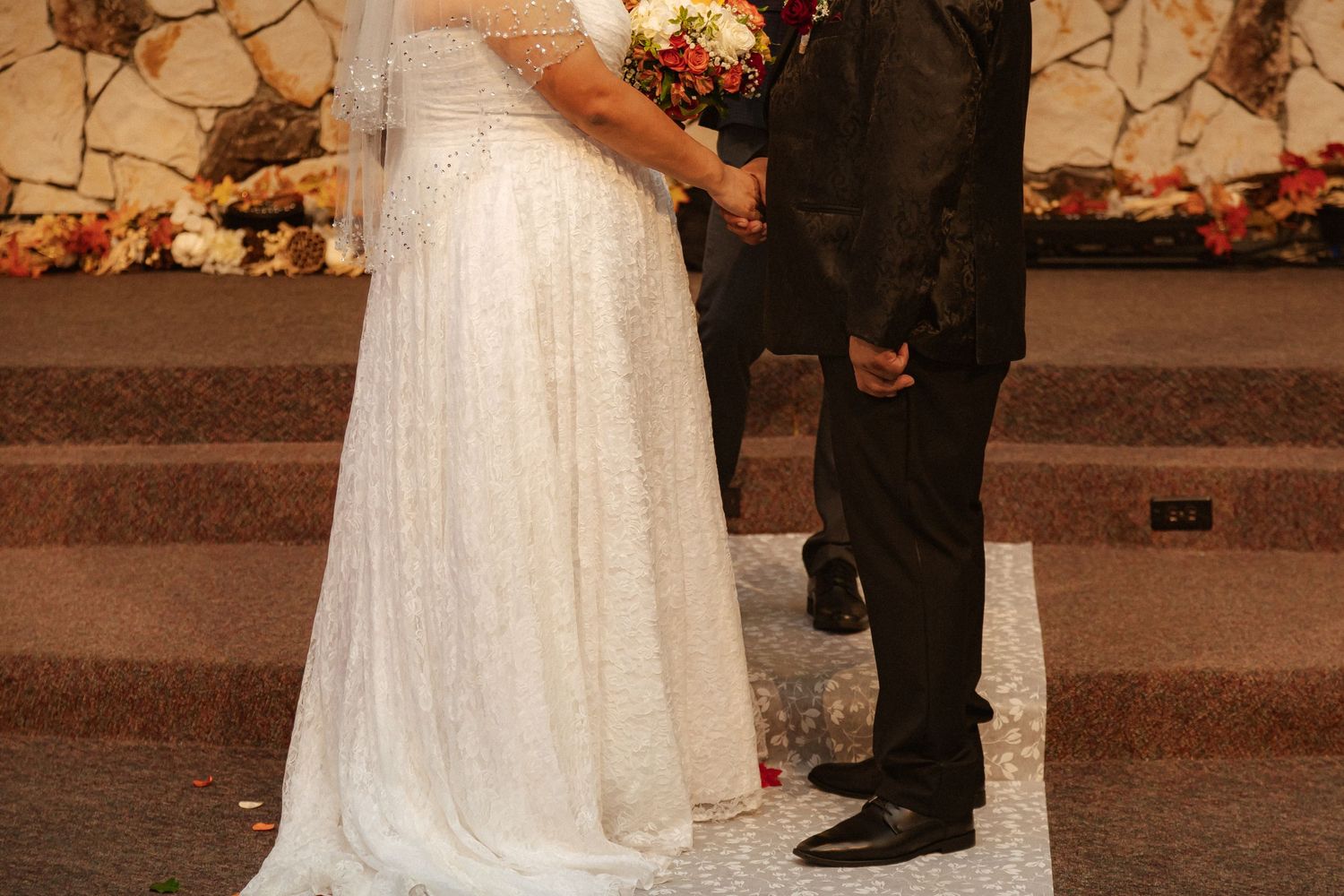 A wedding ceremony taking place in front of a stone wall with a cross and floral decorations.