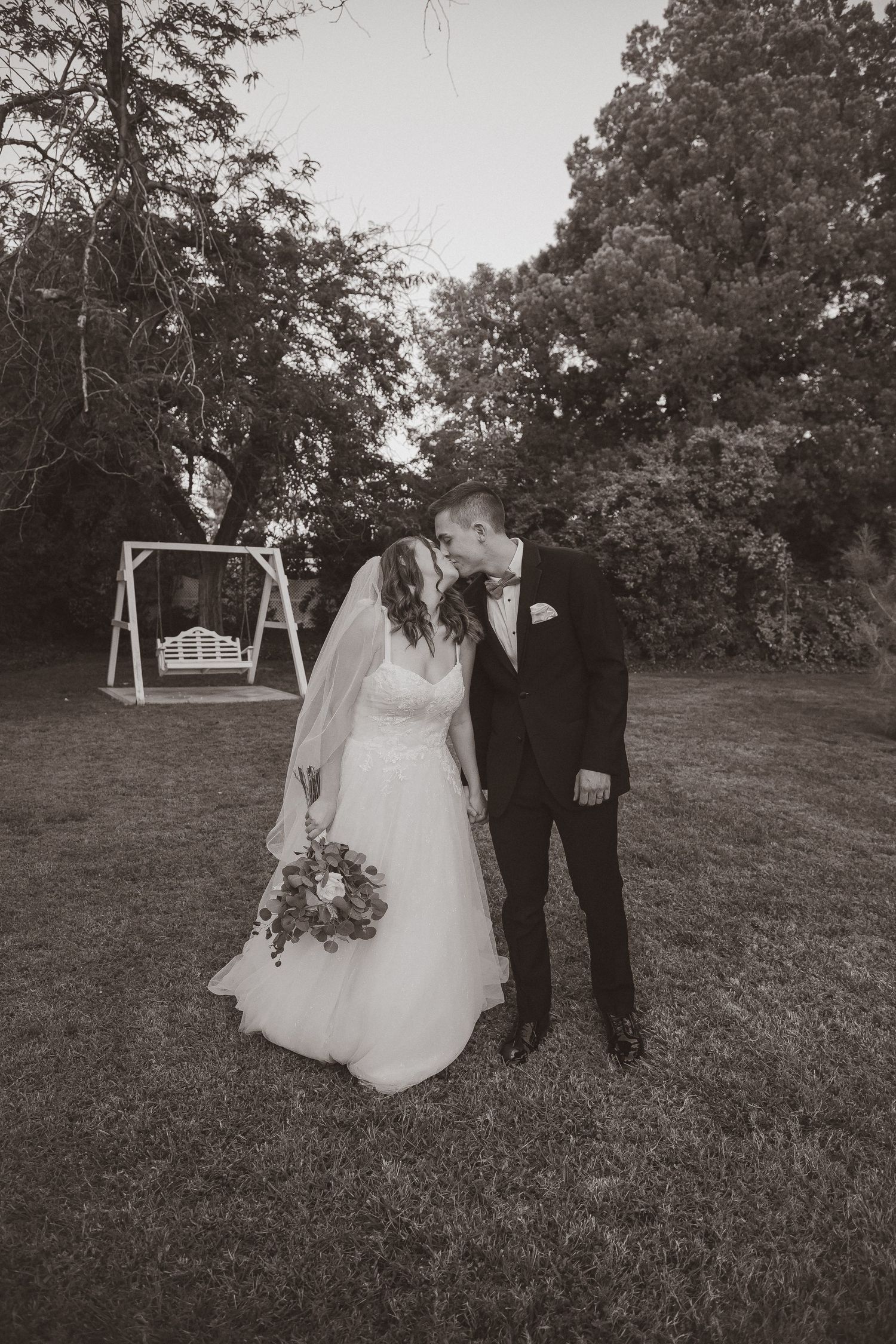Black and white wedding photo with couple standing near a wooden swing set in a garden setting.