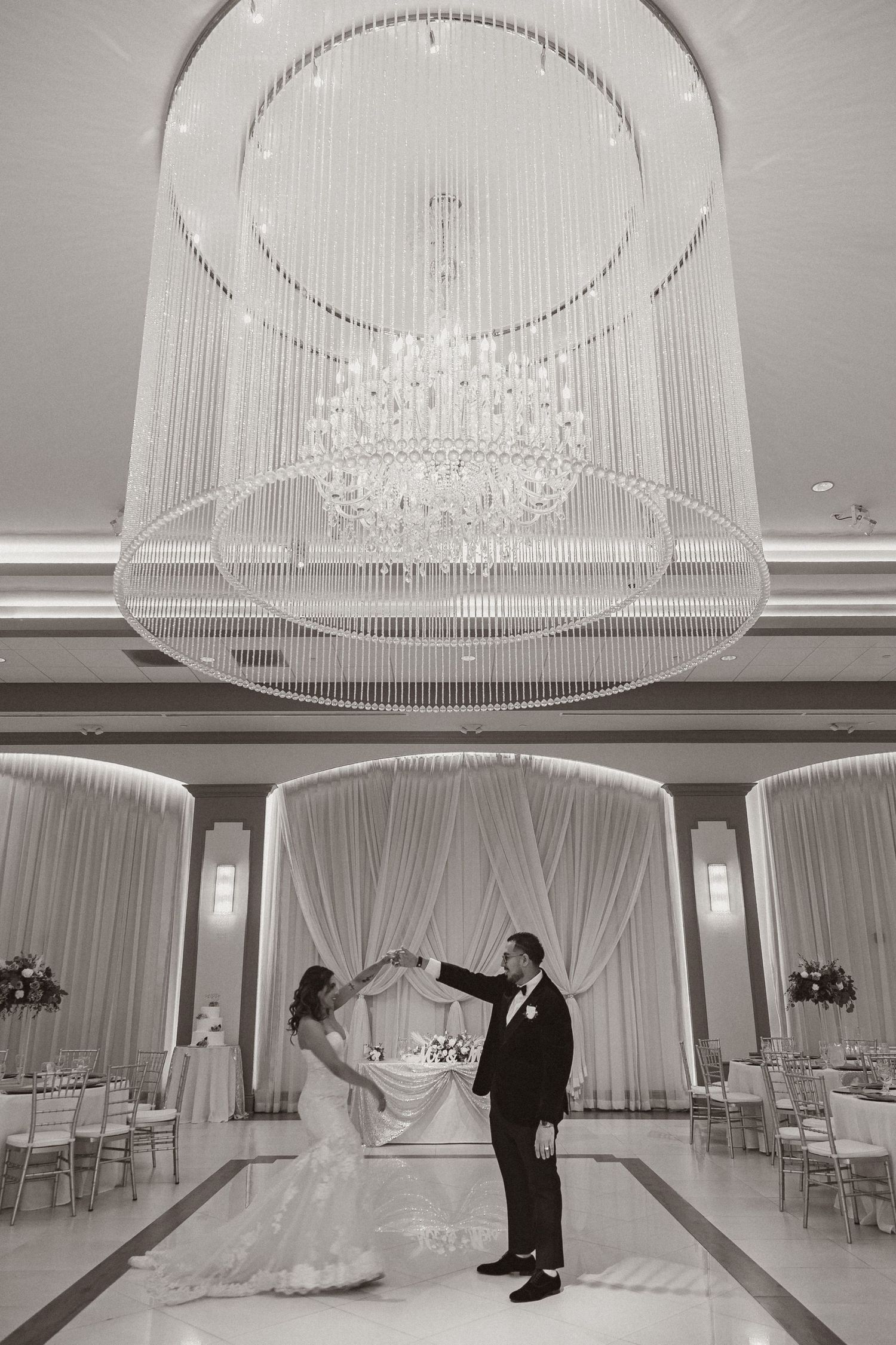 Wedding couple dancing beneath a dramatic crystal chandelier in an elegant white ballroom setting.