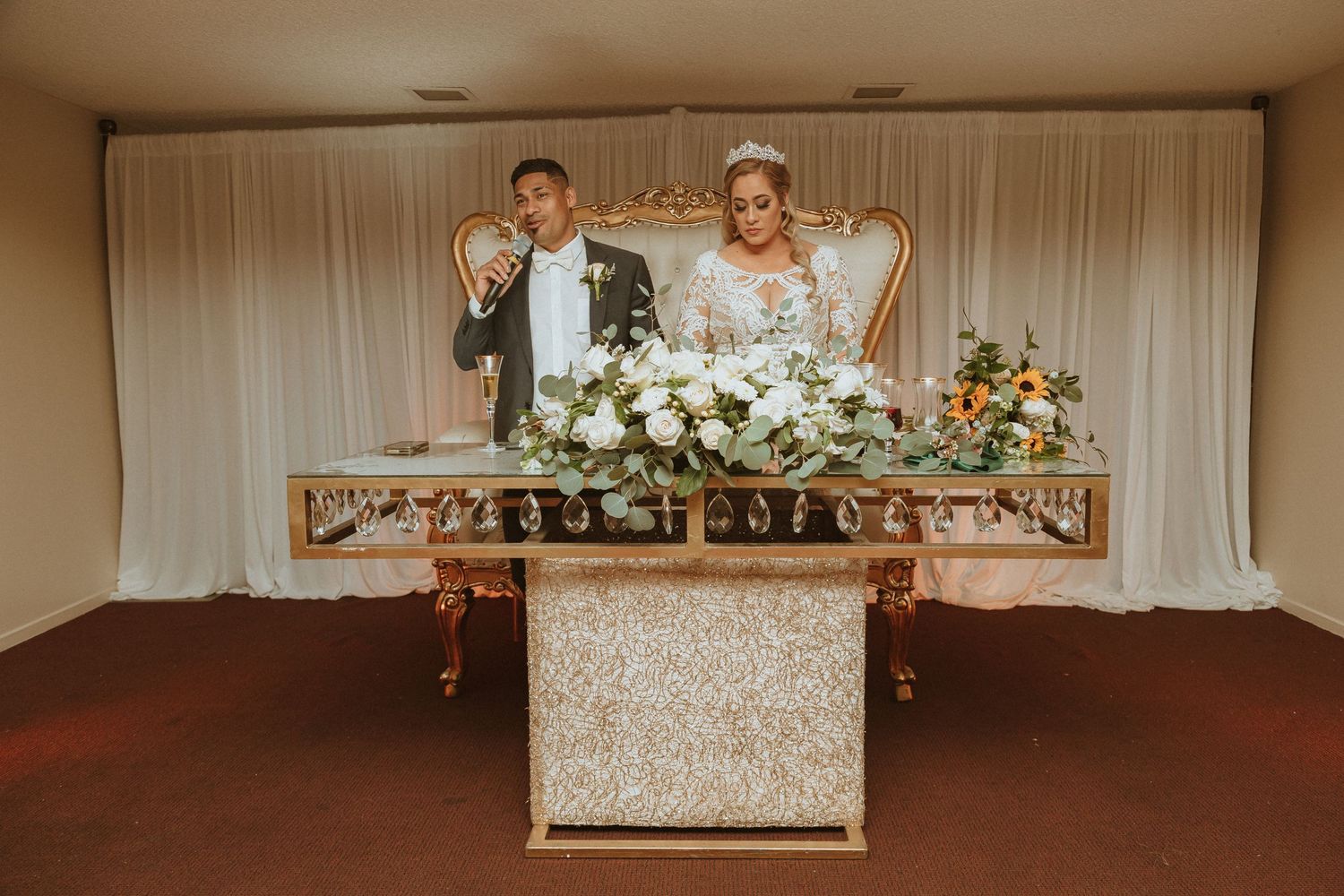 Wedding couple seated at elegant sweetheart table with white floral arrangements and decor.
