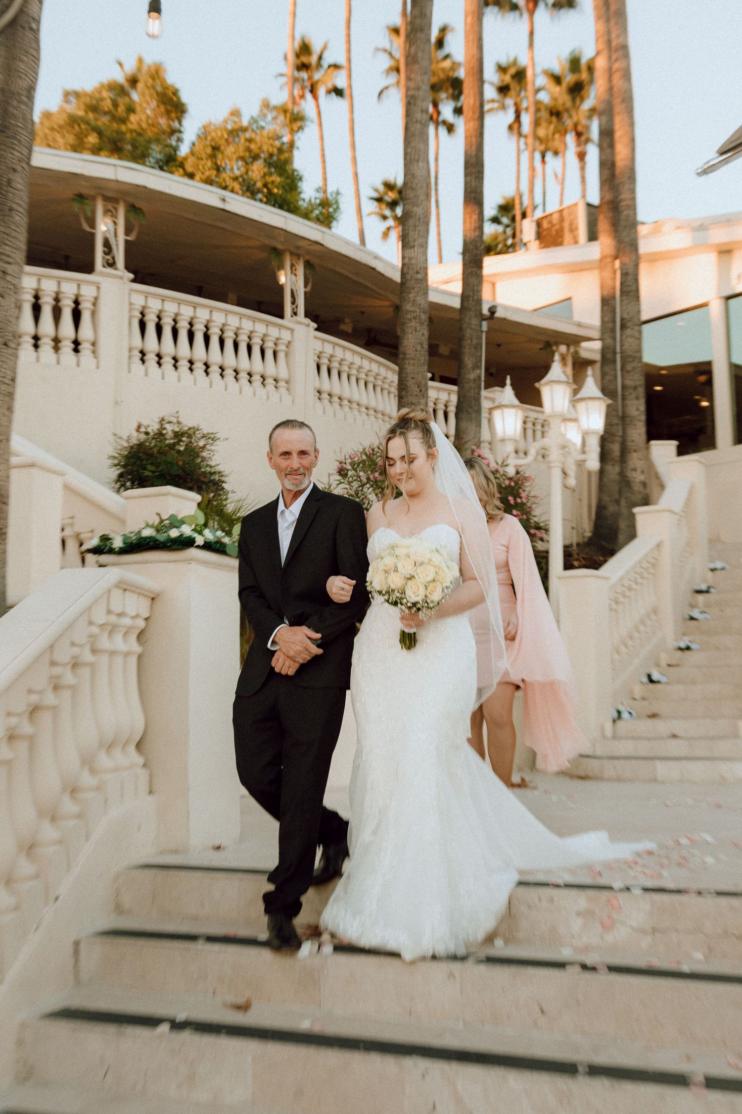 Wedding procession on decorative white staircase with palm trees and string lights.