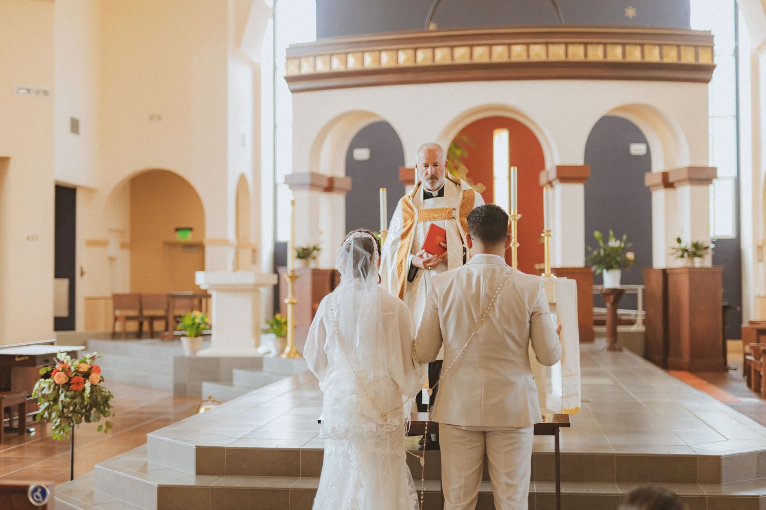 A couple faces the altar during their wedding ceremony in a brightly lit church sanctuary.