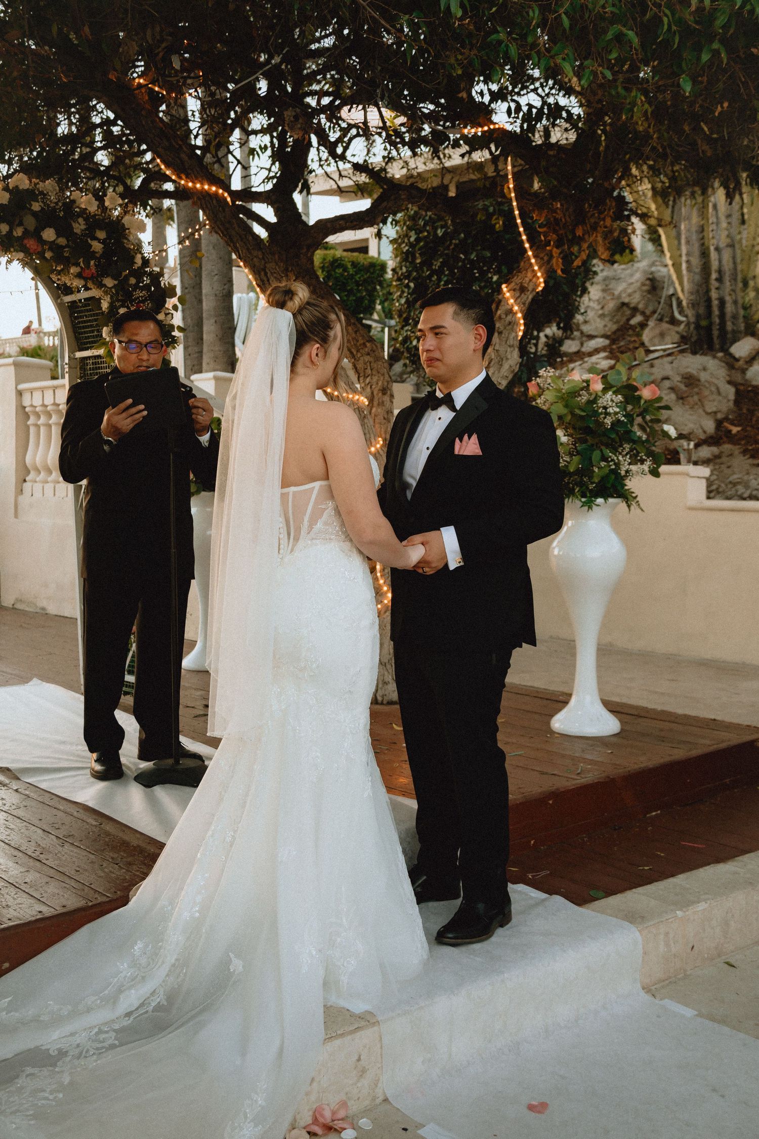 Wedding ceremony under string-lit trees with a couple exchanging vows on a wooden platform.