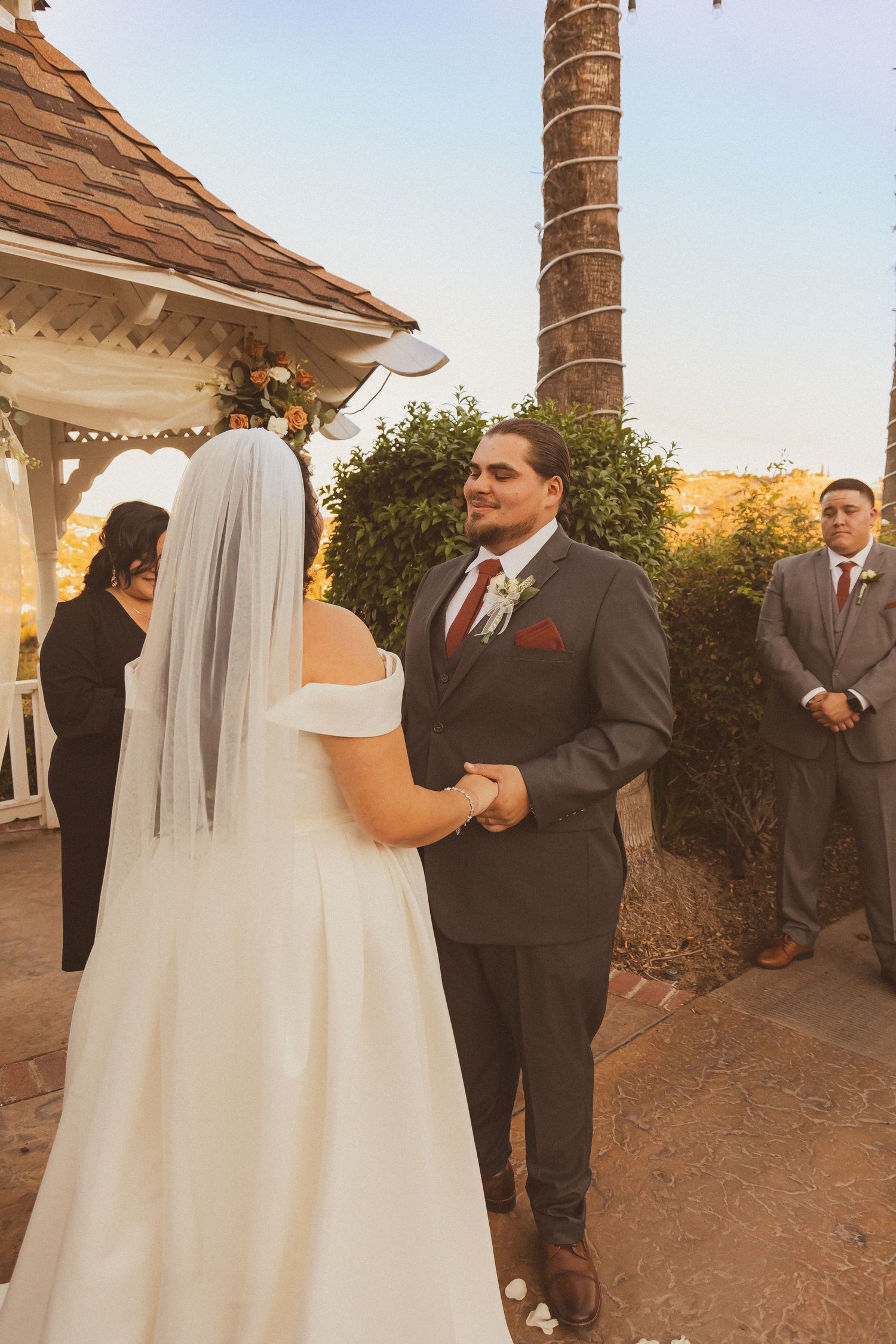 Wedding ceremony takes place under palm trees at sunset with decorated gazebo.