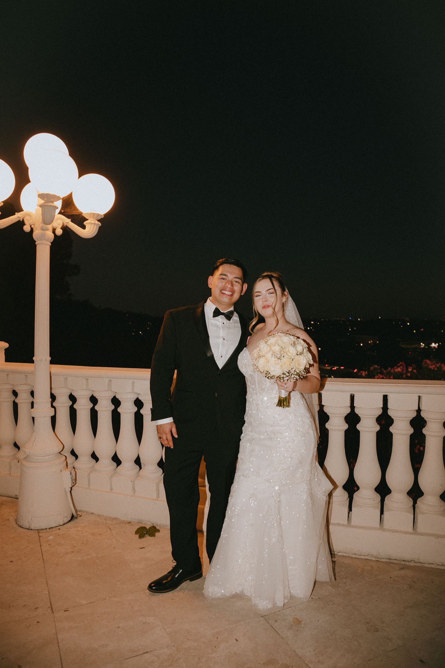 Elegant wedding couple stands on terrace with ornate lighting and city views in background.