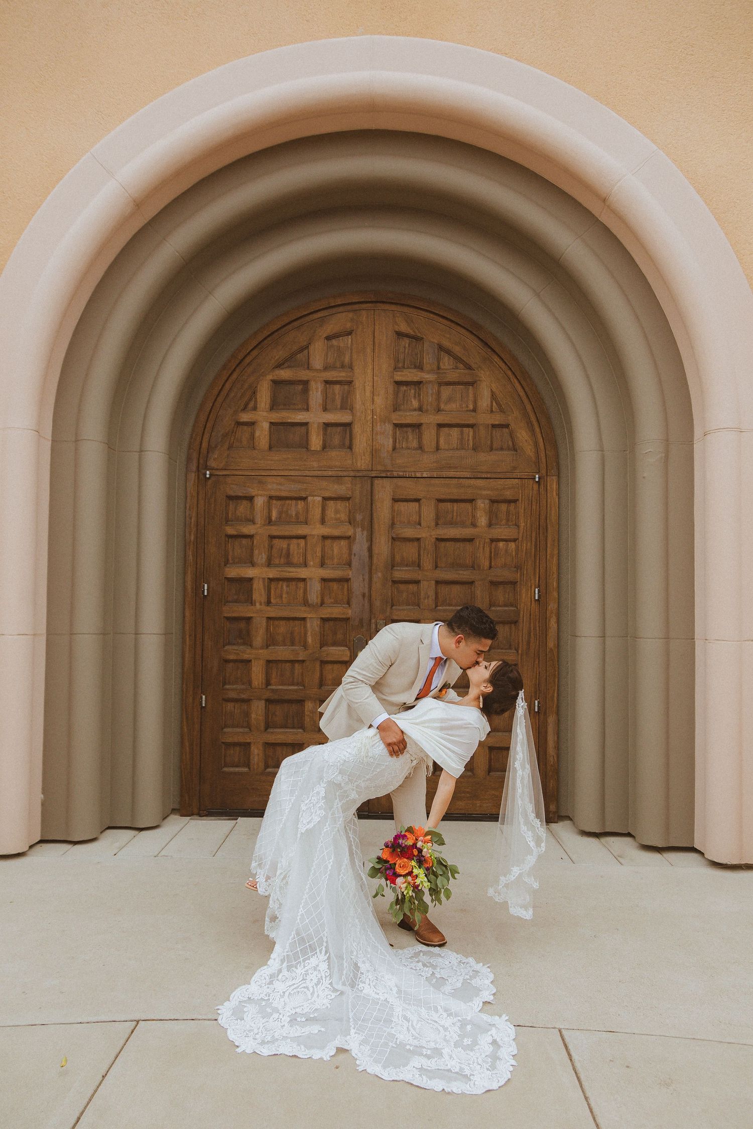 A romantic dip kiss between newlyweds in front of an arched wooden door with the bride holding a bouquet.