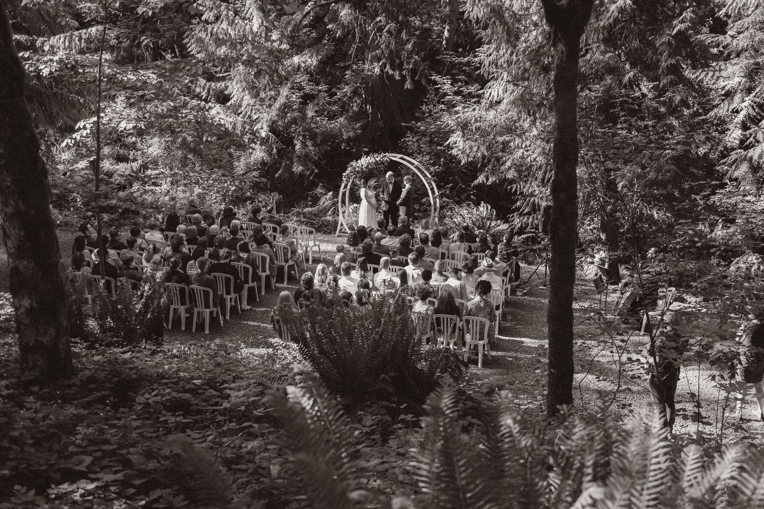 Black and white view of outdoor wedding ceremony with circular arch in forest setting.