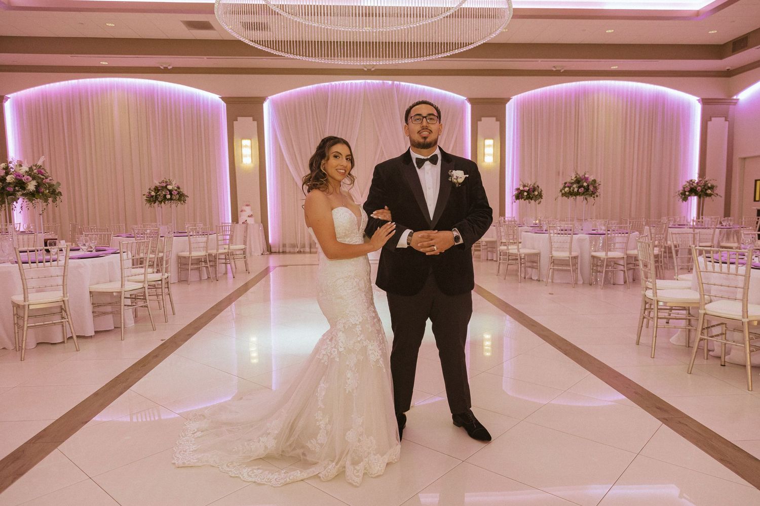Newlyweds posing in elegant ballroom with pink uplighting and white decor.