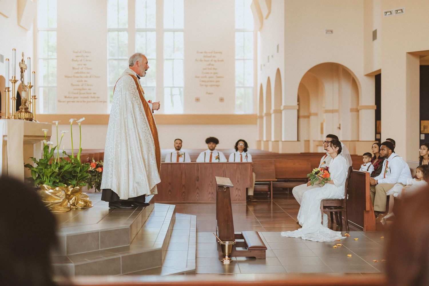 A solemn ceremony takes place in a church with a priest standing on elevated steps near floral arrangements.