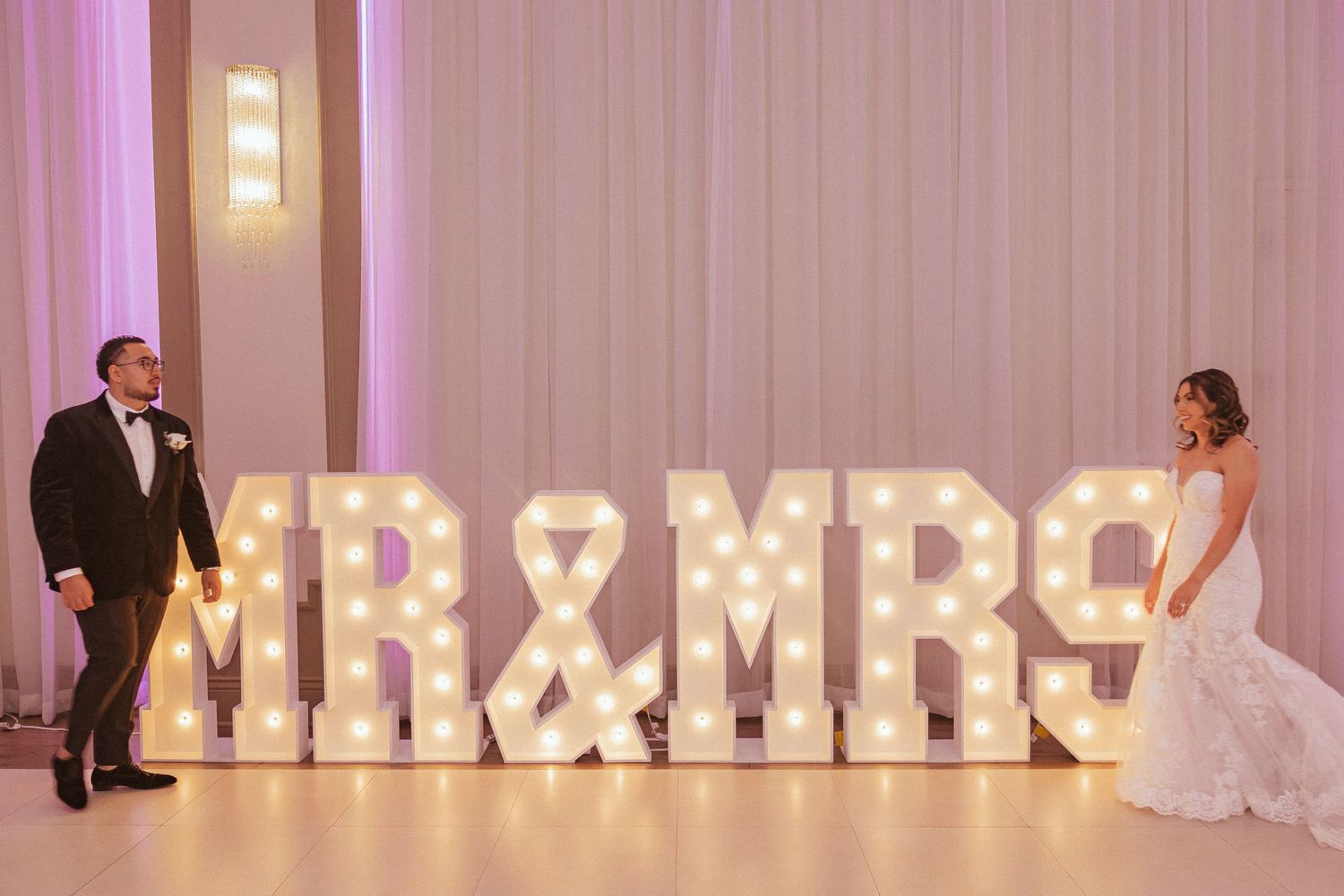 Couple stands on opposite sides of large illuminated MR & MRS sign at wedding reception.