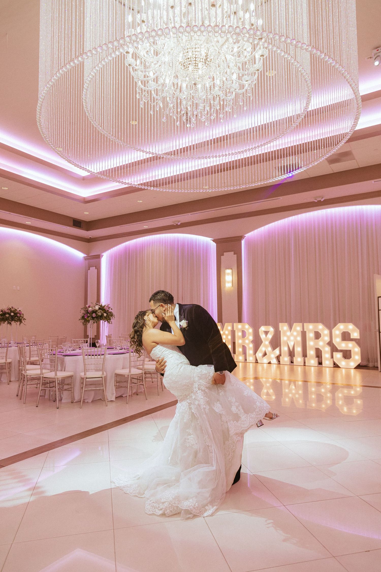 Dramatic dip kiss between newlyweds in an elegant pink-lit ballroom with illuminated MR & MRS sign.