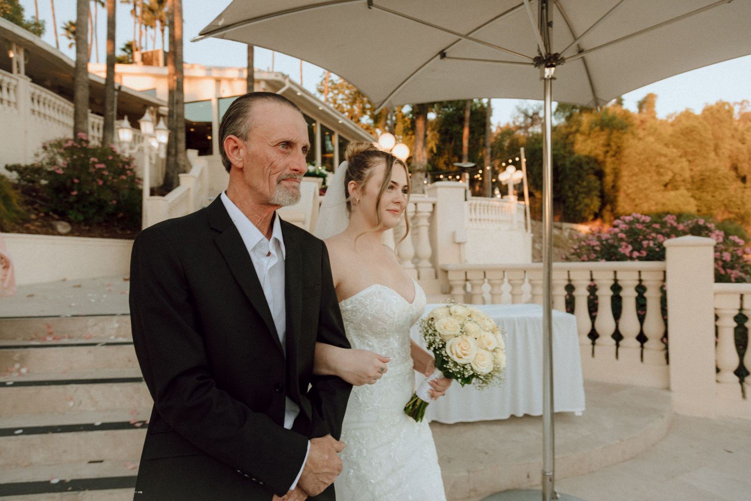 Outdoor wedding ceremony on decorative white stairs with palm trees and parasols in golden hour lighting.