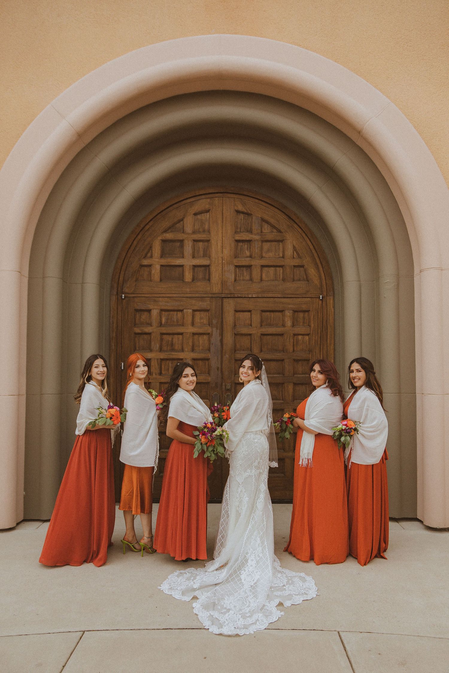 Bridal party in flowing orange dresses and white wraps pose with bride in lace gown against stone archway.