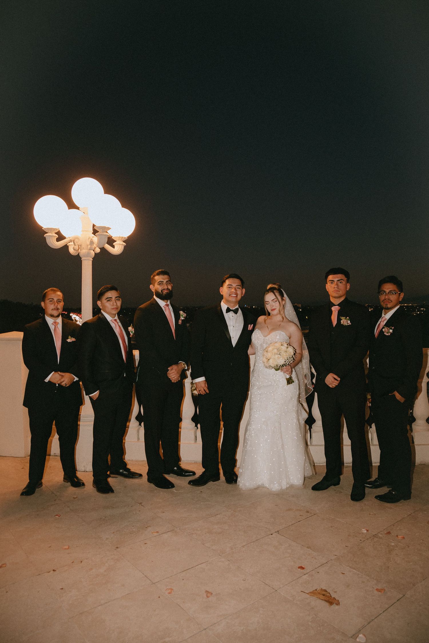 Wedding group poses outdoors under glowing vintage street lamp during evening celebration.