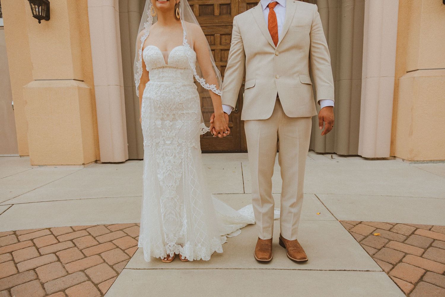 Couple in elegant wedding attire holding hands on a brick paved walkway.