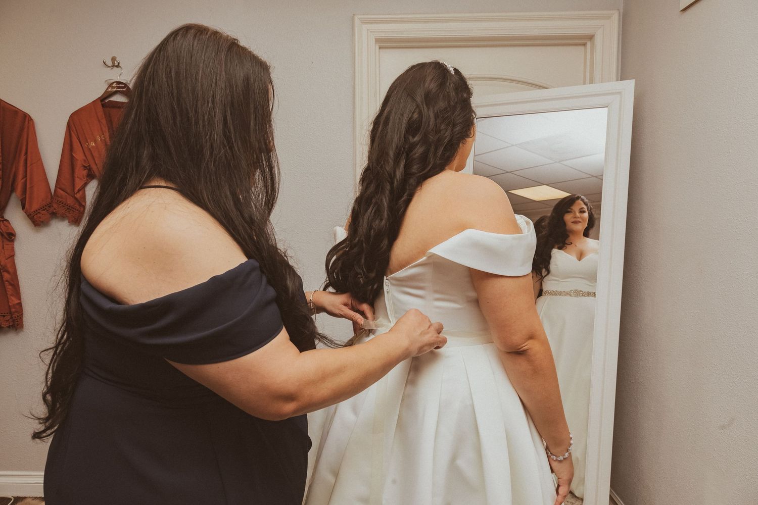 A helper assists with final wedding dress adjustments while looking in a mirror.