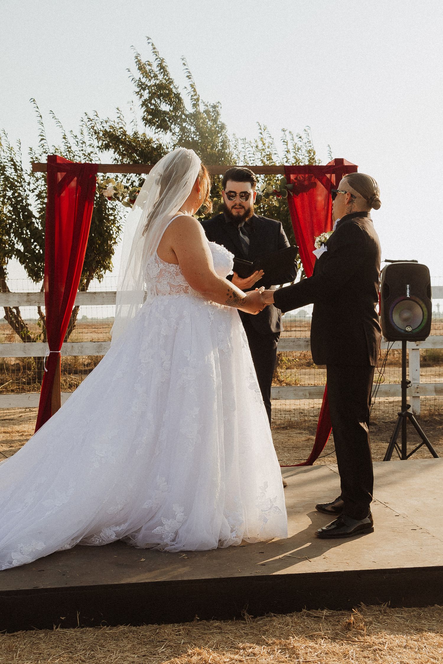 An outdoor wedding ceremony under a red-draped arch with a speaker system visible.
