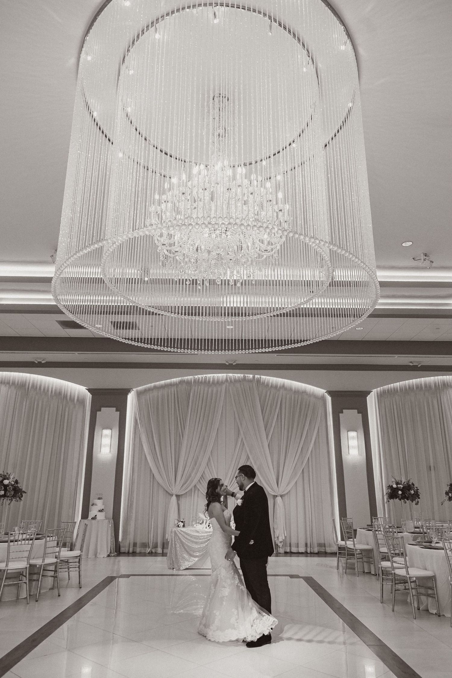 Newlyweds share an intimate dance beneath a grand crystal chandelier in a beautifully decorated ballroom.